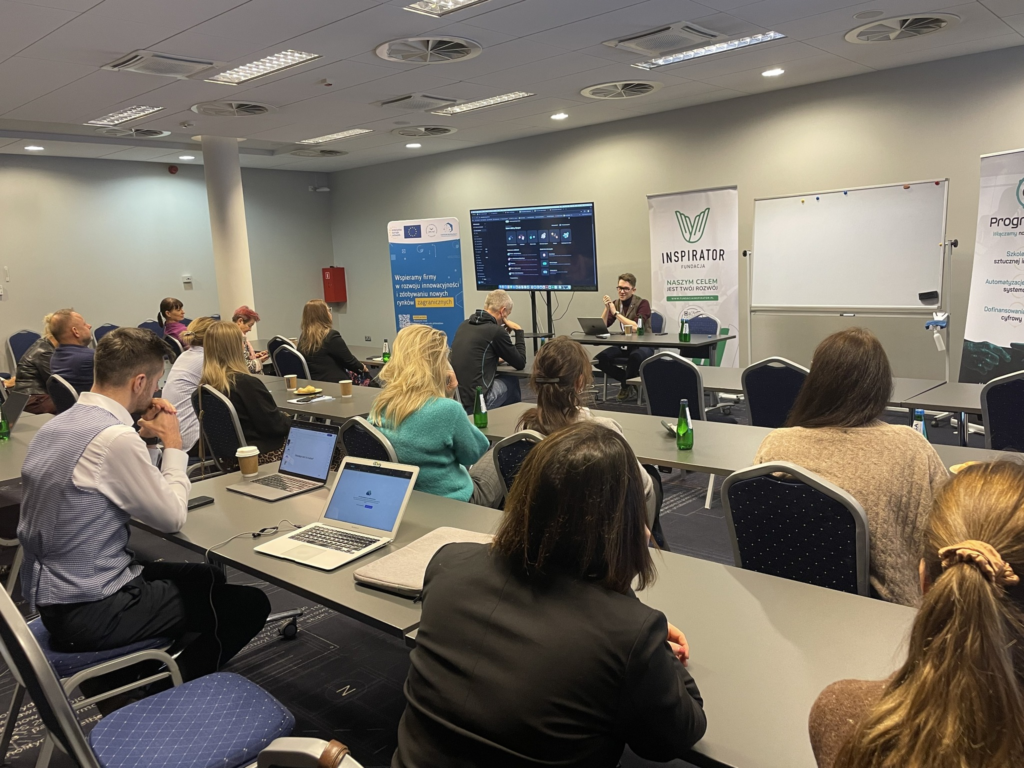 Zdjęcie z warsztatu: prezentowany jest ekran z Elleven labs - platformy, która pozwala na budowanie agentów AI w biurach obsługi klientów. Na sali jest dużo osób, które siedzą plecami do kamery. Na zdjeciu jest prowadzący Kacper Rzosiński